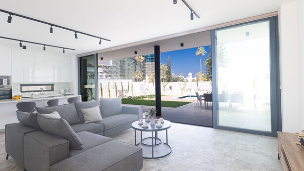 A modern living room with a grey sectional sofa, a round coffee table, and a large sliding glass door leading to an outdoor area with a pool and palm trees. - Protaras Aqua Pearl Villa AQ05 by Interhome (Protaras)