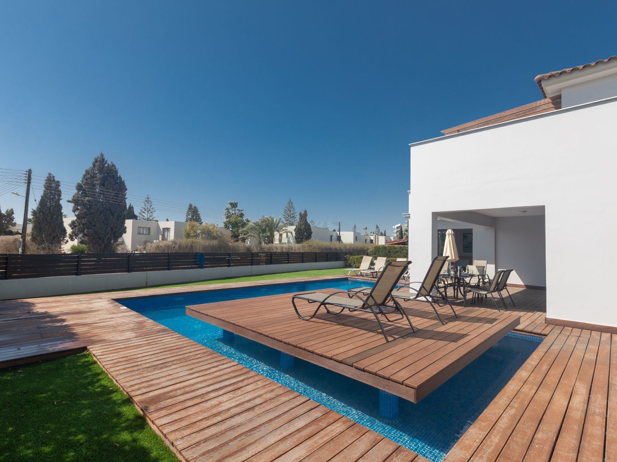 Ein moderner Poolbereich im Freien mit Holzdeck, Liegestühlen und einem weißen Gebäude im Hintergrund.