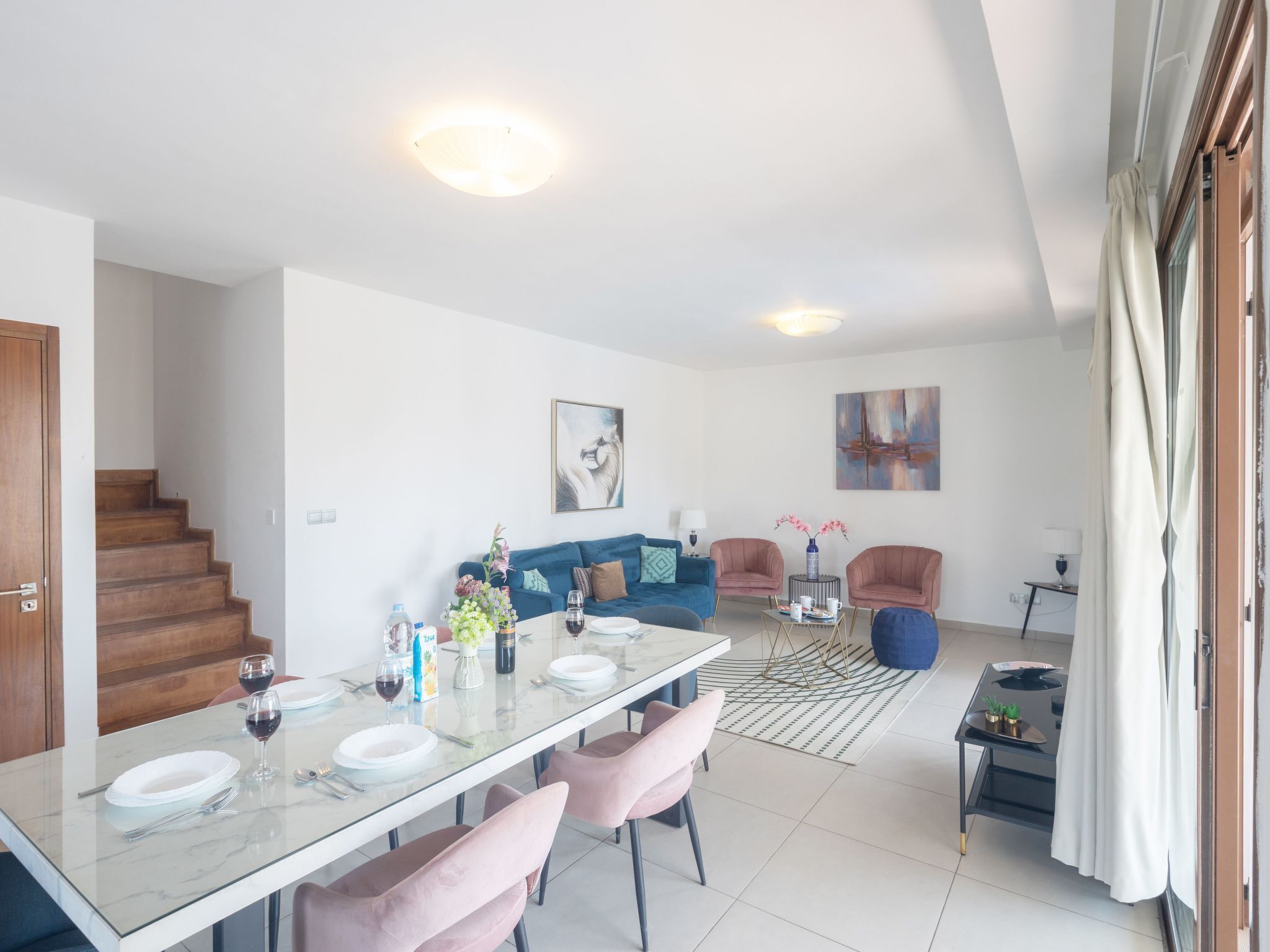 A modern living room with a dining area, featuring a staircase, a blue sofa, a coffee table, and a painting on the wall.