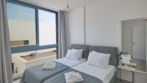 Een moderne hotelkamer met een groot raam, een bed met twee kussens, een nachtkastje met een lamp en een kleine tafel met een ventilator.