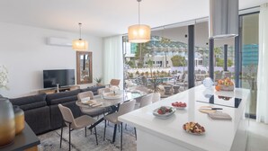 Un moderno salón con un sofá, una mesa de comedor y sillas. Hay un televisor, un ventanal grande y vistas a un edificio.