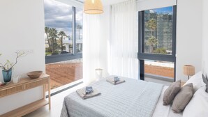 Un dormitorio moderno con una cama grande, una mesita de noche con lámpara, y vistas a palmeras y edificios a través de amplias ventanas.