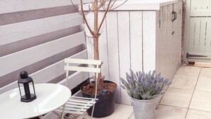 Un pati amb taula i cadires blanques, una planta de lavanda en test i una paret de pedra.