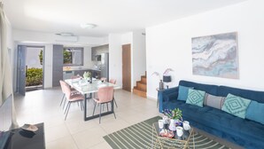 Een moderne woonkamer met een blauwe bank, een eettafel met roze stoelen, en een keuken op de achtergrond.