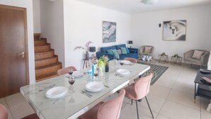 Een moderne woonkamer met een eethoek, met een glazen tafel, roze stoelen, een blauwe bank en een trap.