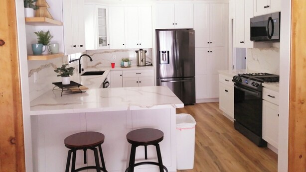A modern kitchen with white cabinets, a stainless steel refrigerator, a microwave, and a stove. There are two bar stools and a trash bin. - Bright beach-city home — newly renovated. Only 3 mi. from SoCal beaches!   (Torrance)