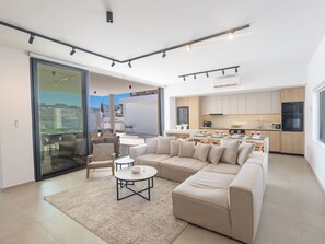 En moderne stue med en stor beige hjørnesofa, et rundt sofabord og en glasdør, der fører til en udendørs terrasse.