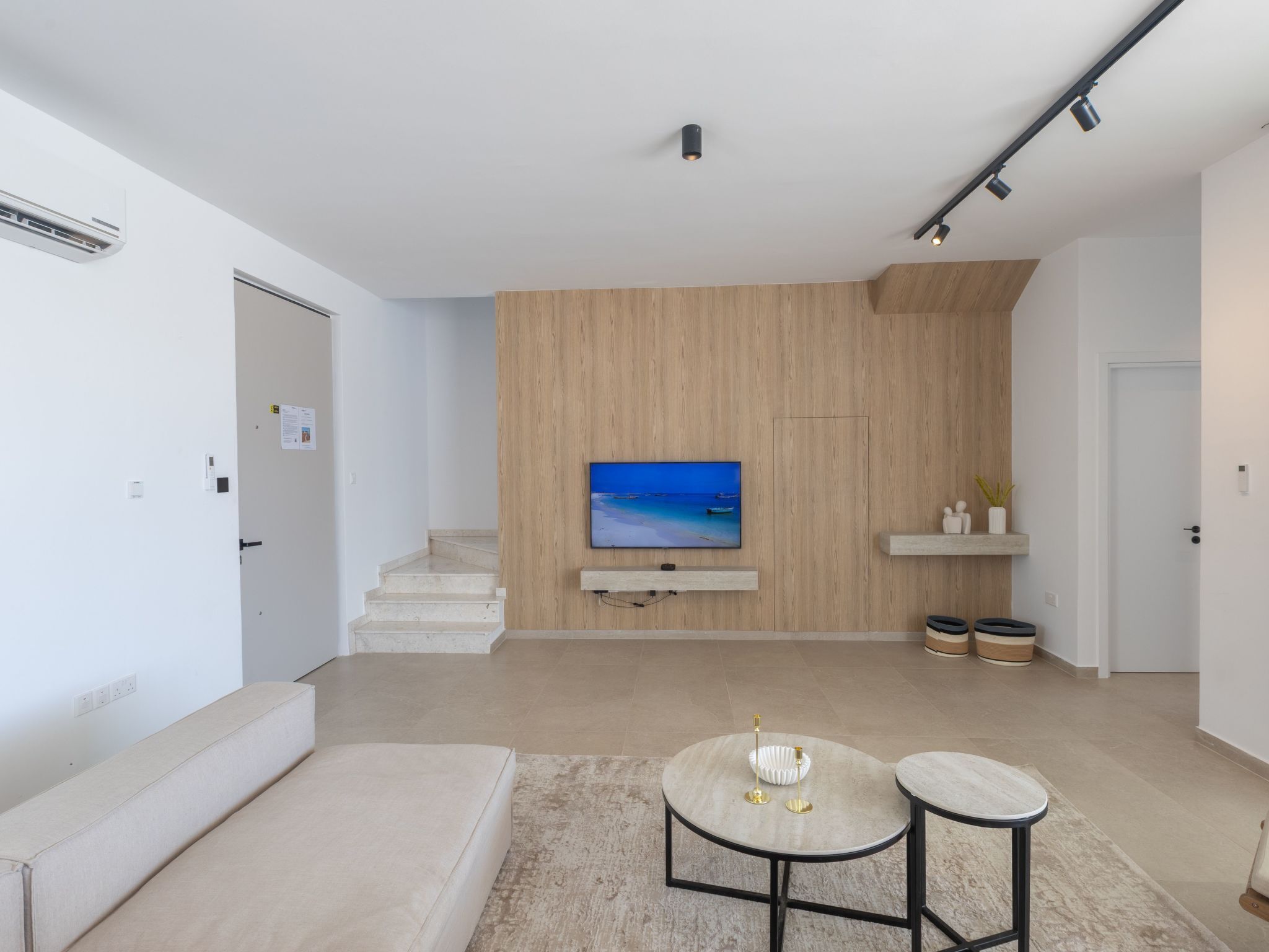 Un salon moderne avec un téléviseur à écran plat fixé à un mur en bois, un escalier menant à un autre niveau et une décoration minimaliste agrémentée d’un tapis et de deux petites tables.