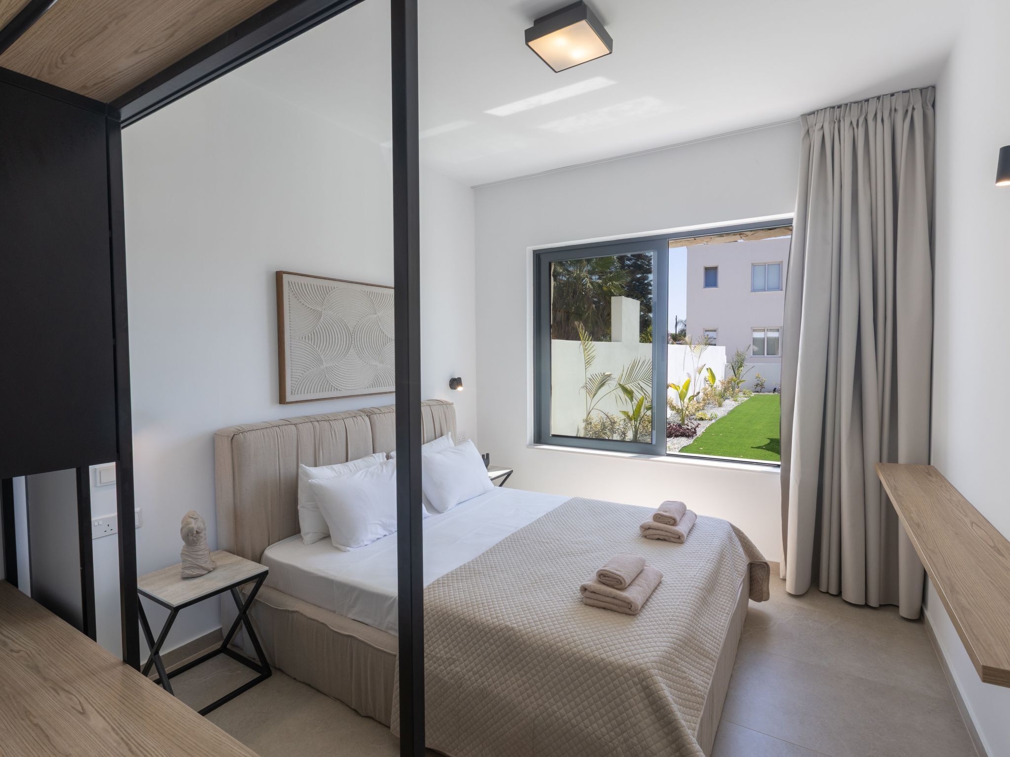 Ein modernes Schlafzimmer mit einem großen Bett, einem Nachttisch, einem Fenster mit Blick ins Grüne und einer Deckenleuchte.