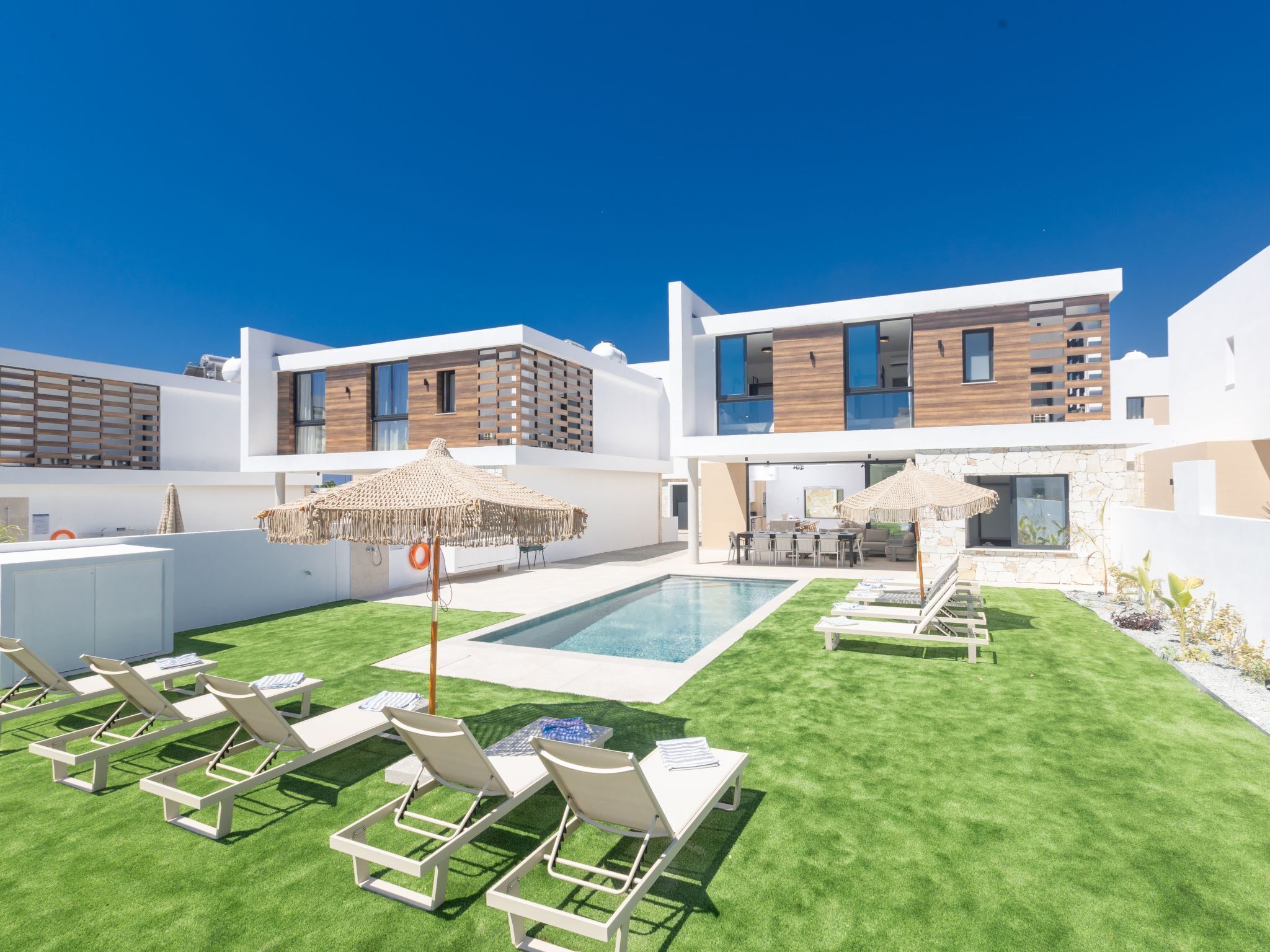 Un espace piscine extérieur moderne, doté de chaises longues et de parasols, entouré d’un bâtiment à deux étages dont la façade combine des éléments en bois et des finitions blanches.