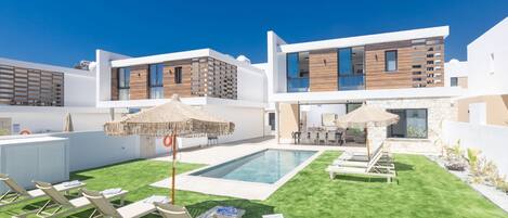 Un espace piscine extérieur moderne, doté de chaises longues et de parasols, entouré d’un bâtiment à deux étages dont la façade combine des éléments en bois et des finitions blanches.