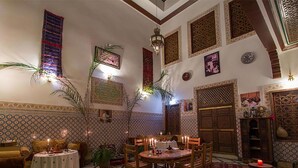 Un intérieur marocain traditionnel, avec un sol à carreaux, des murs décorés de motifs et un coin repas où une table est dressée pour quatre personnes.