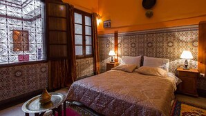 Une chambre à coucher avec un grand lit, deux tables de chevet avec des lampes, une tête de lit à motifs, une fenêtre ornée de barreaux en fer décoratifs et une petite table avec une théière.