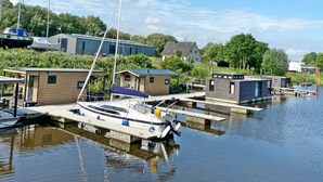 A marina with several boats docked, a small wooden building, and a modern house in the background. - Hausboot KHT 2 Lacet mit Elektrokamin im Salon, 1 Schlafraum, Küche, Duschbad m by Interhome (Wilhelmshaven)