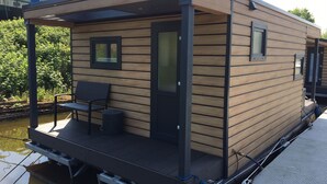 A modern floating house with a deck, a bench, and a small table. - Hausboot KHT 2 Lacet mit Elektrokamin im Salon, 1 Schlafraum, Küche, Duschbad m by Interhome (Wilhelmshaven)