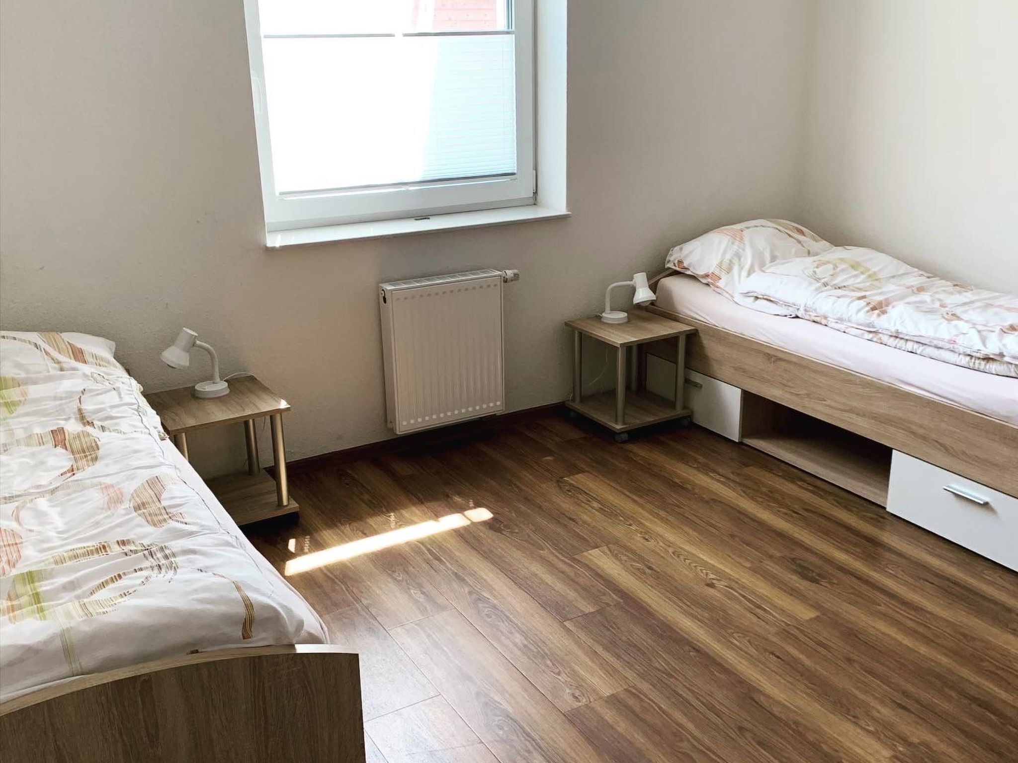 A bedroom with two beds, a window, a radiator, and a bedside table with a lamp.