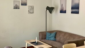 Olohuoneessa on ruskea sohva, pieni puinen pöytä ja lattiavalaisin. Seinällä on neljä kehystettyä kuvaa.