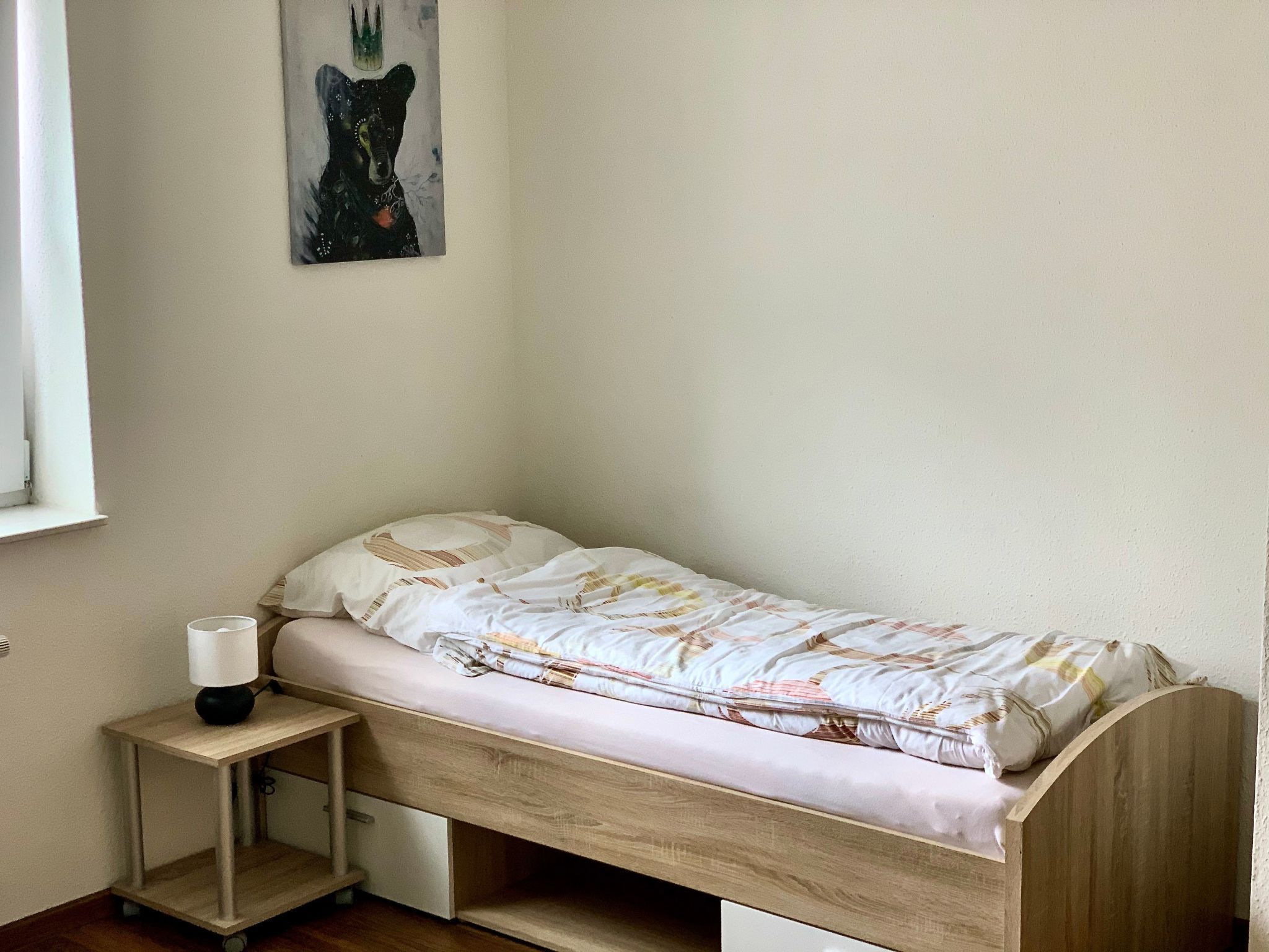 A small, single-bed room with a wooden headboard, a bedside table with a lamp, and a wall-mounted painting of a bear.