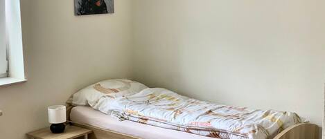 A small, single-bed room with a wooden headboard, a bedside table with a lamp, and a wall-mounted painting of a bear.