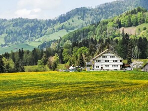 Un edificio a due piani con tetto spiovente, circondato da un campo verde e dolci colline ricoperte di alberi.