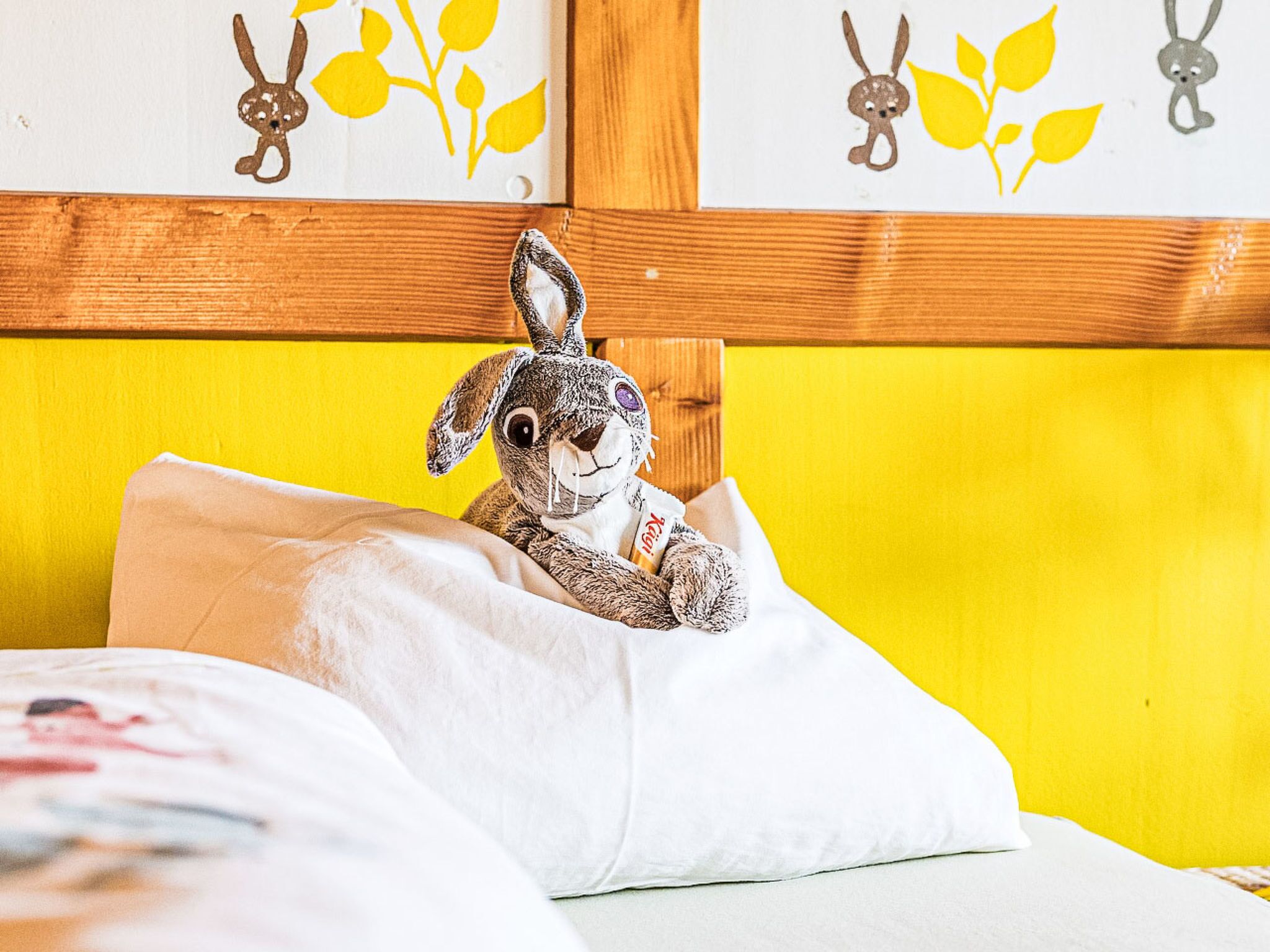 Ein Bett mit Stoff-Hasen, Kissen und einem hölzernen Kopfteil, an einer gelben Wand.