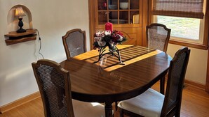 Uma sala de jantar com uma mesa de madeira, quatro cadeiras, um candelabro, uma janela com persianas e um armário de madeira com louças.