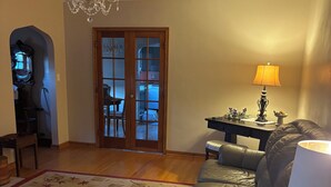 Uma sala de estar com um candelabro, um sofá, uma mesa redonda e uma porta de madeira que dá para outro cômodo.