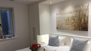 Quarto com cama, almofadas, mesa de cabeceira, janela e uma obra de arte na parede.