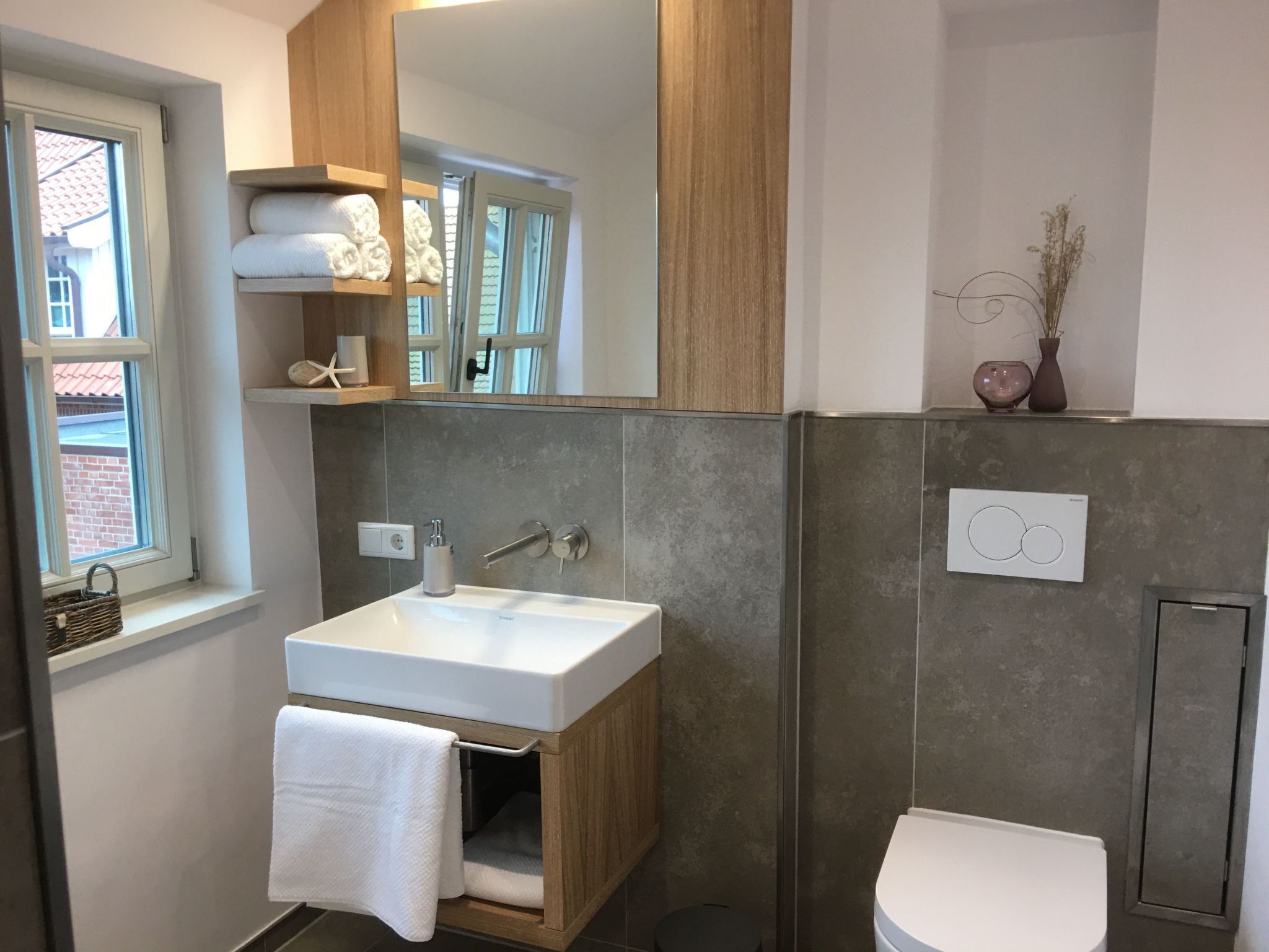 Un baño moderno con un mueble de tocador de madera, un lavabo blanco y un inodoro. Hay una ventana que ofrece vistas a un edificio de ladrillo.