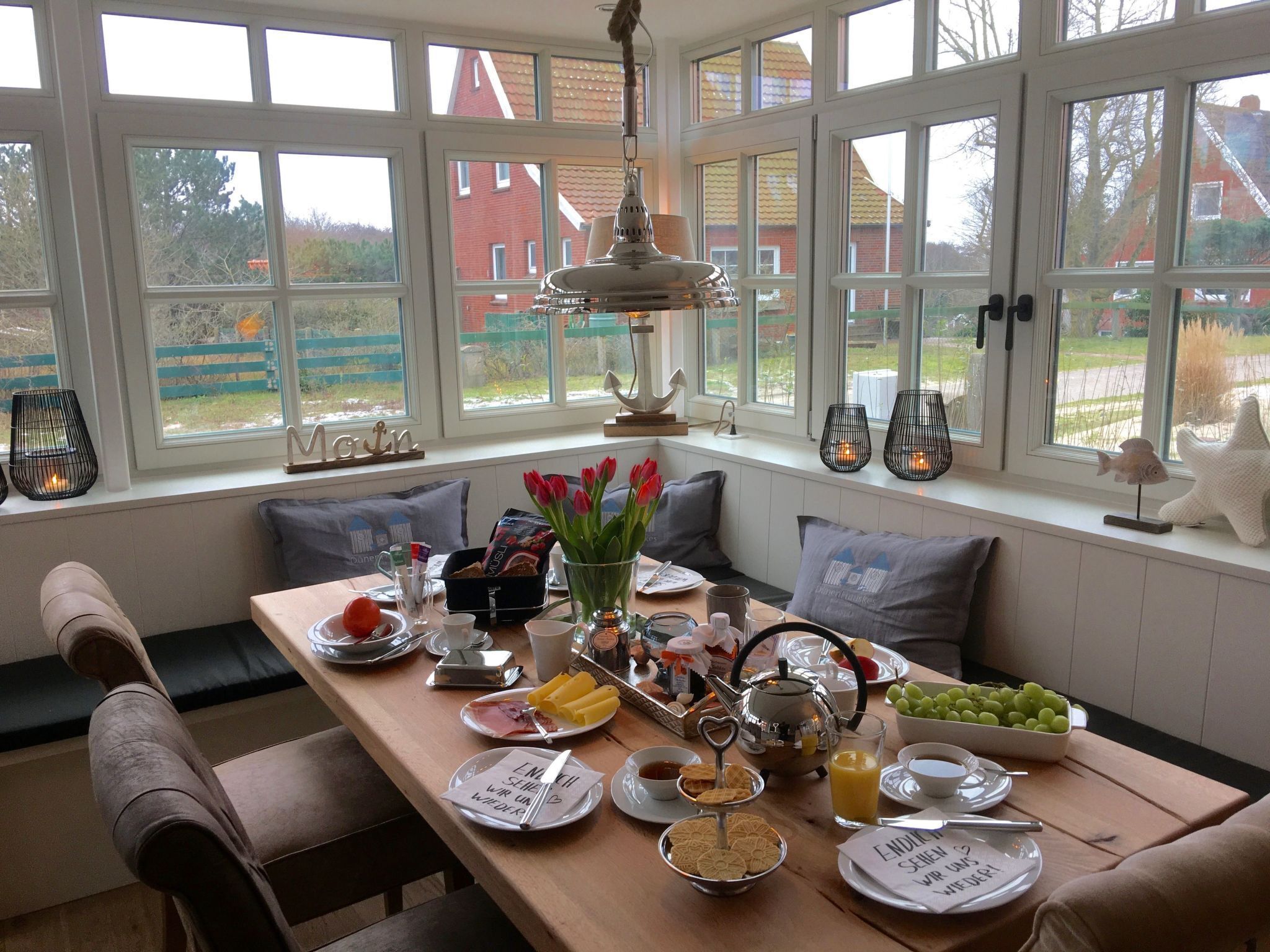 A breakfast setup with a table, breakfast items, and a view of a red house through large windows.