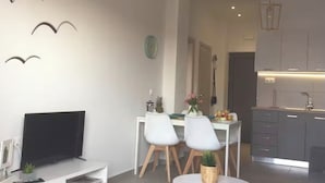 Ein modernes Wohnzimmer mit Flachbildfernseher, eine Essecke mit zwei Stühlen, einem kleinen runden Tisch und eine Küche mit grauen Schränken und einer Arbeitsplatte.