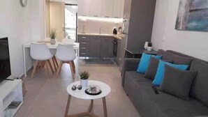Ein modernes Wohnzimmer mit einem grauen Sofa und einem kleinen runden Tisch. Es gibt eine Essecke mit einem weißen Tisch und Stühlen. Im Hintergrund ist eine Küche mit modernen Geräten erkennbar.