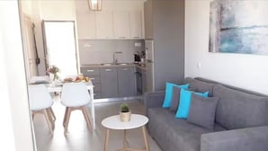 Ein modernes Wohnzimmer mit grauem Sofa und blauen Kissen, einem kleinen runden Tisch mit Pflanze sowie einem Essbereich mit weißen Stühlen und Tisch.