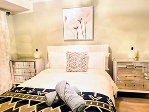 A neatly made bed with a patterned blanket, a decorative pillow, and a towel on the side. There is a framed artwork above the bed, a bedside table with a lamp, and a dresser with a mirror.