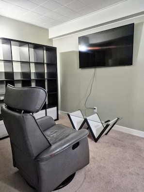 A room with a gray leather chair, a glass coffee table, a flat-screen TV mounted on the wall, and a black shelving unit.