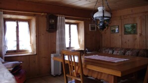 Interior d'una cabana de fusta amb una taula de dinar, cadires, una banca, una làmpada penjant i un rellotge a la paret.