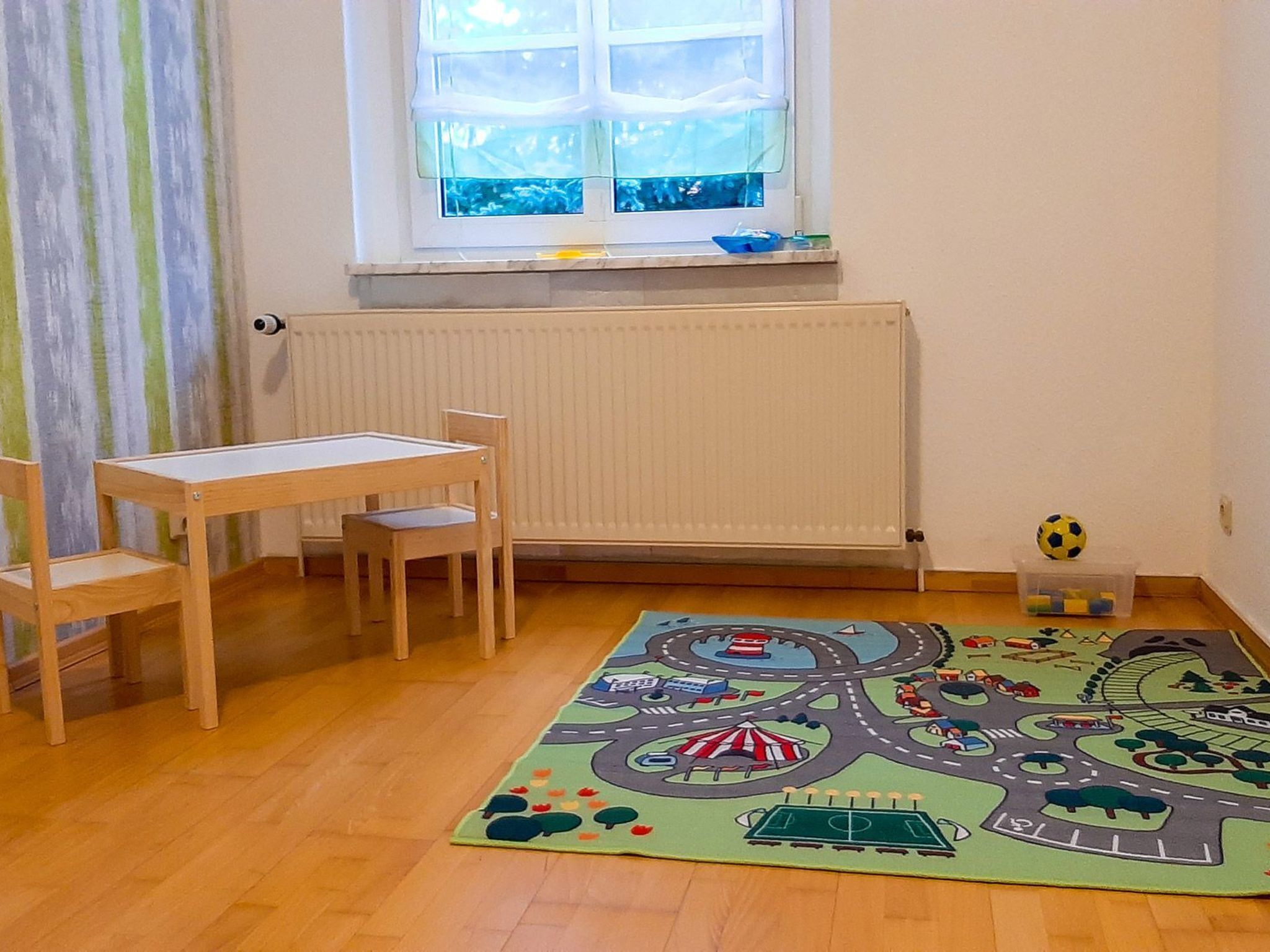 Una habitación con una mesa y sillas de madera, una ventana con persianas, un radiador y una alfombra de juego colorida con un diseño de carretera y vehículos.
