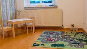 Une pièce avec une table et des chaises en bois, une fenêtre avec des stores, un radiateur et un tapis de jeu coloré représentant une route et des véhicules.