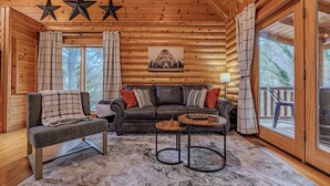 Acogedor interior de cabaña de madera con sofá, sillón y mesa de centro, paredes de madera, ventanas amplias y un adorno con forma de estrella.