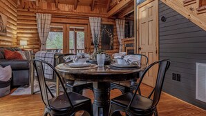 Interior de una cabaña de madera con una mesa de comedor preparada para cuatro personas, un sofá y una escalera que conduce a un nivel superior.