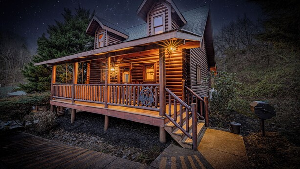 Una cabaña de troncos con terraza de madera y porche cubierto, rodeada de árboles y un cielo nocturno estrellado.