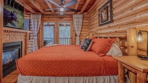 Un dormitorio tipo cabaña de madera con una cama grande, chimenea, ventilador de techo y vistas al exterior.