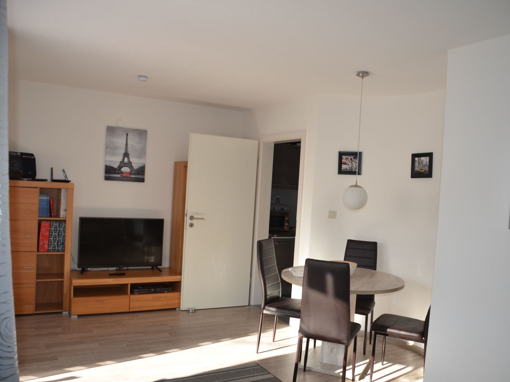 Una sala moderna con televisor de pantalla plana, mueble de entretenimiento de madera, mesa de comedor con sillas y una puerta que da a una cocina.