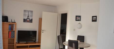 Una sala moderna con televisor de pantalla plana, mueble de entretenimiento de madera, mesa de comedor con sillas y una puerta que da a una cocina.