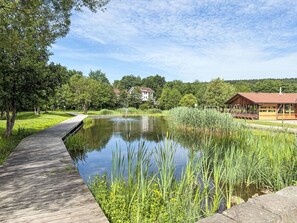 Ein hölzerner Steg führt zu einem Teich, im Hintergrund sind ein Pavillon und ein Haus zu sehen.