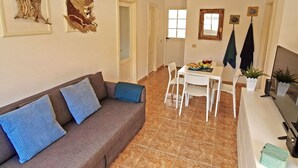 A living room with a brown sofa, white chairs, a dining table with a fruit bowl and wine glasses, and a TV on a stand. - Casa Cristof by Interhome (Costa Calma)