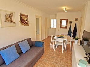 Ein Wohnzimmer mit einem braunen Sofa, weißen Stühlen, einem Esstisch mit Obstschale und Weingläsern sowie einem Fernseher auf einem Lowboard.