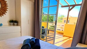 A hotel room with a bed, a sun-shaped wall decoration, a small plant, and a view of a yellow building through a glass door. - Casa Cristof by Interhome (Costa Calma)