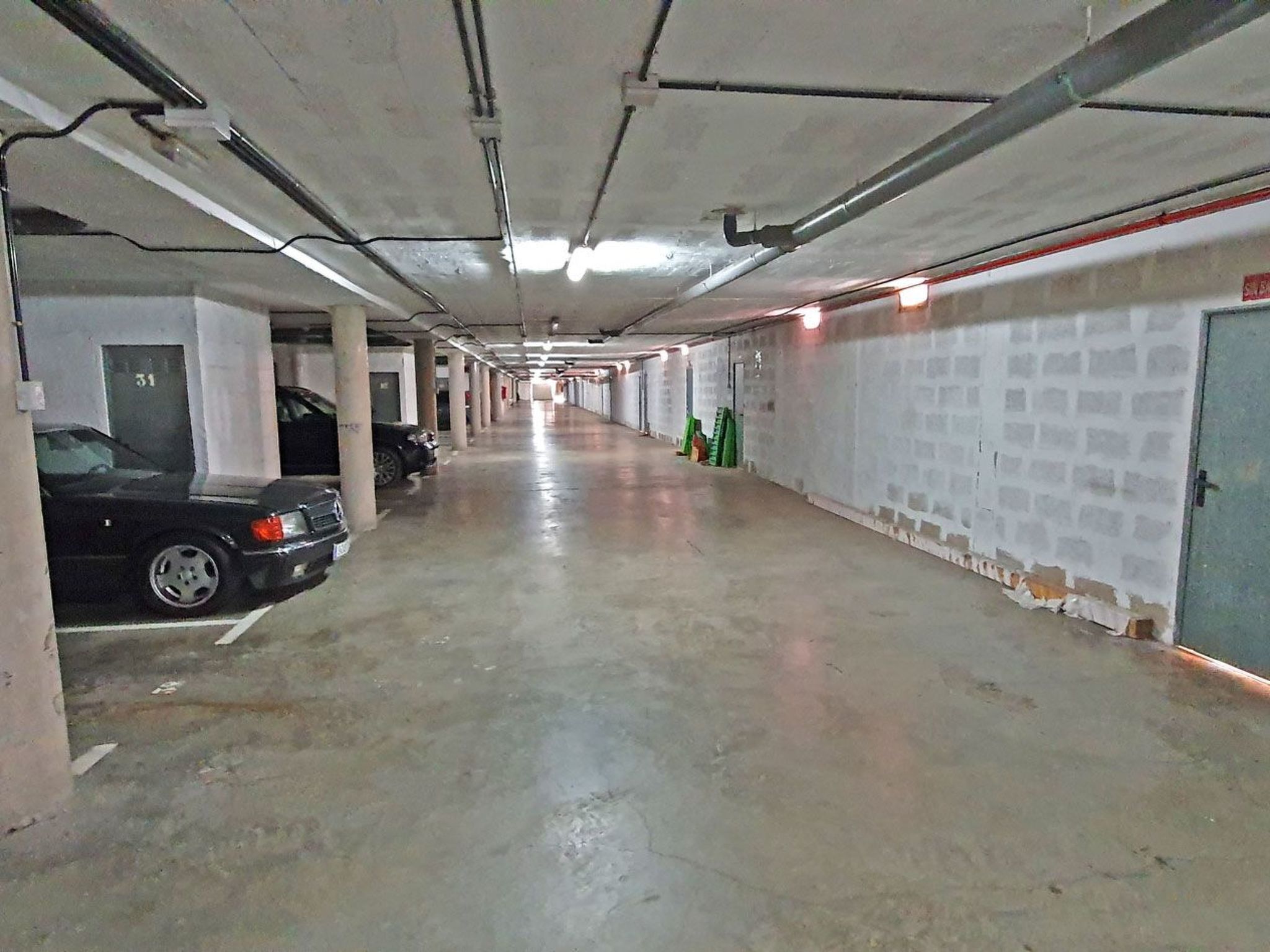Eine lange, schmale Tiefgarage mit Betonböden und -wänden, in der sich mehrere Autos befinden und Rohre an der Decke sichtbar sind.