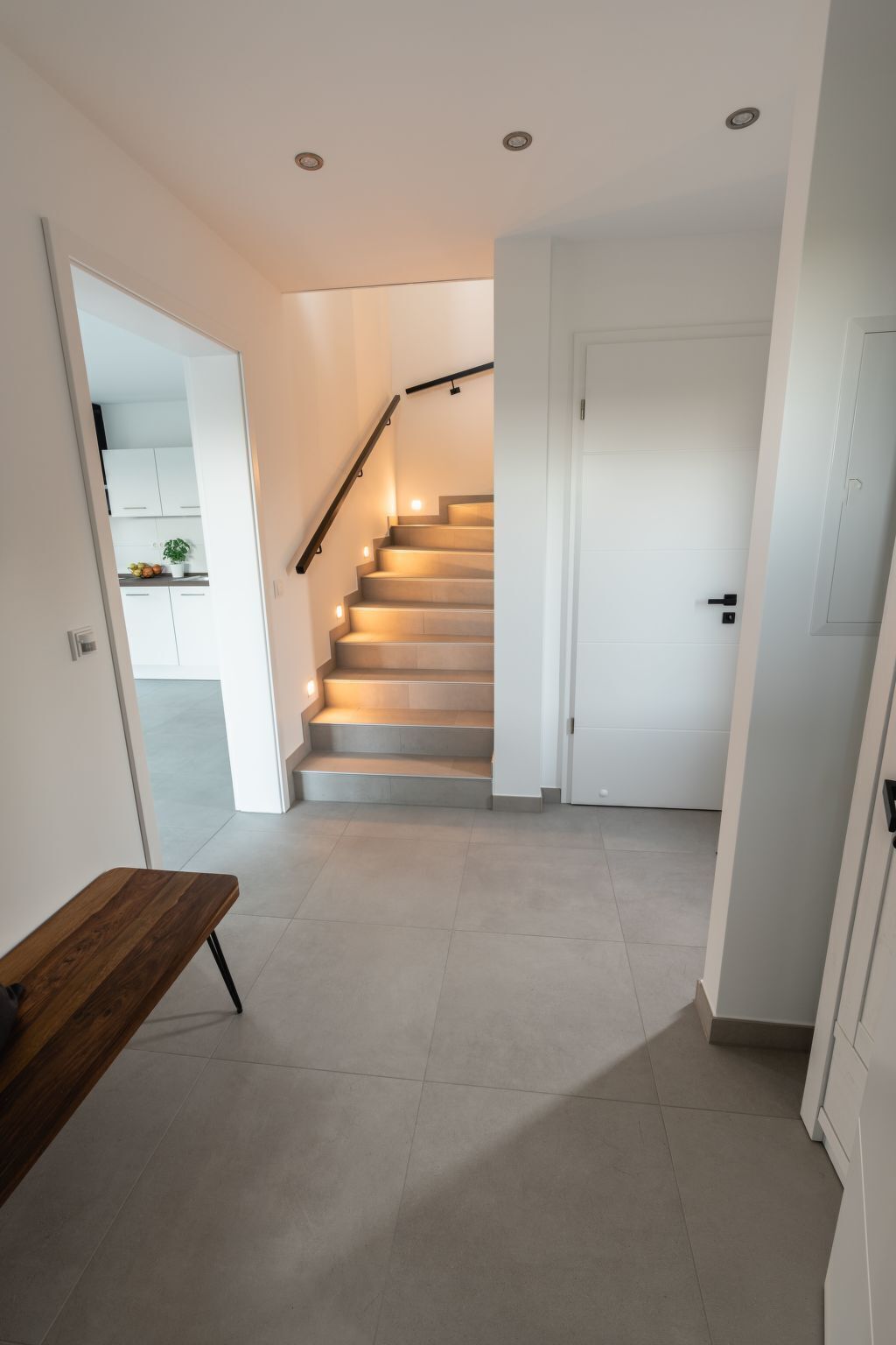 A modern interior with a staircase leading to a second floor, a wooden bench, and a white door.